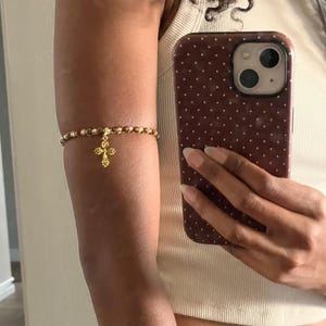 K&ouml;nnte beinhalten: Ein Oberarmband bestehend aus einer Reihe kleiner, runder, goldfarbener Metallperlen. Ein goldfarbener Kreuzanh&auml;nger mit einem strukturierten, blumenartigen Design h&auml;ngt in der Mitte des Perlenbandes und liegt am Oberarm an.