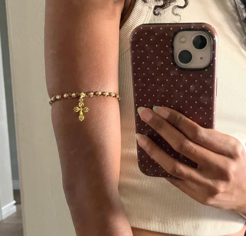 K&ouml;nnte beinhalten: Ein Oberarmband bestehend aus einer Reihe kleiner, runder, goldfarbener Metallperlen. Ein goldfarbener Kreuzanh&auml;nger mit einem strukturierten, blumenartigen Design h&auml;ngt in der Mitte des Perlenbandes und liegt am Oberarm an.
