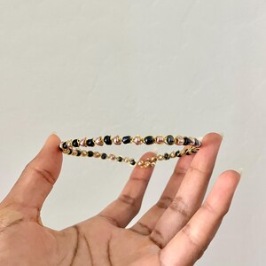 K&ouml;nnte beinhalten: Ein kreisf&ouml;rmiges Armband aus d&uuml;nnem, goldfarbenem Draht, der um kleine, glatte, kugelf&ouml;rmige Perlen in Schwarz und metallischem Ros&eacute;gold gewickelt ist und ein durchgehendes, filigranes und leichtes Banddesign bildet.