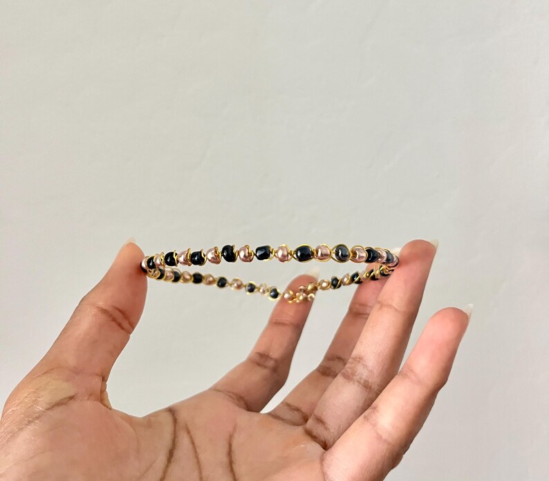 K&ouml;nnte beinhalten: Ein kreisf&ouml;rmiges Armband aus d&uuml;nnem, goldfarbenem Draht, der um kleine, glatte, kugelf&ouml;rmige Perlen in Schwarz und metallischem Ros&eacute;gold gewickelt ist und ein durchgehendes, filigranes und leichtes Banddesign bildet.