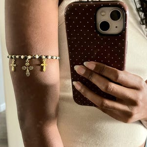 K&ouml;nnte beinhalten: Ein Armband bestehend aus einer Reihe abwechselnd wei&szlig;er und metallisch dunkelgrauer Perlen. Drei goldfarbene Kreuzanh&auml;nger h&auml;ngen an der Mitte des Perlenstrangs, wobei ein gr&ouml;&szlig;eres, verziertes Kreuz zwischen zwei kleineren, schlichten Kreuzen sitzt.