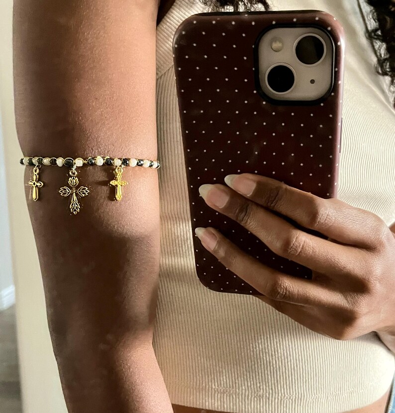 K&ouml;nnte beinhalten: Ein Armband bestehend aus einer Reihe abwechselnd wei&szlig;er und metallisch dunkelgrauer Perlen. Drei goldfarbene Kreuzanh&auml;nger h&auml;ngen an der Mitte des Perlenstrangs, wobei ein gr&ouml;&szlig;eres, verziertes Kreuz zwischen zwei kleineren, schlichten Kreuzen sitzt.