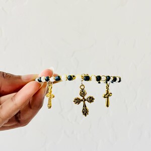 K&ouml;nnte beinhalten: Ein Perlenarmband mit abwechselnd schwarzen und wei&szlig;en Perlen auf einem goldfarbenen Draht. Das Armband ist mit drei goldfarbenen Metallkreuz-Anh&auml;ngern verziert, darunter ein gr&ouml;&szlig;eres zentrales Kreuz mit Blattmuster und zwei kleinere Kreuze.