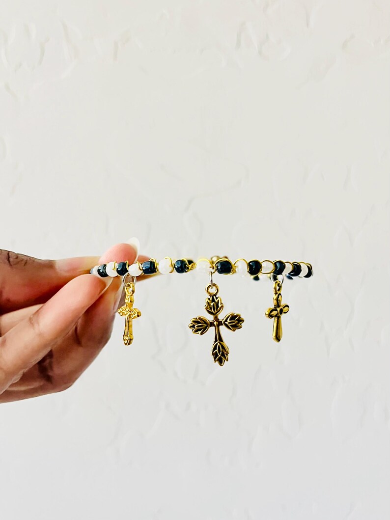 K&ouml;nnte beinhalten: Ein Perlenarmband mit abwechselnd schwarzen und wei&szlig;en Perlen auf einem goldfarbenen Draht. Das Armband ist mit drei goldfarbenen Metallkreuz-Anh&auml;ngern verziert, darunter ein gr&ouml;&szlig;eres zentrales Kreuz mit Blattmuster und zwei kleinere Kreuze.
