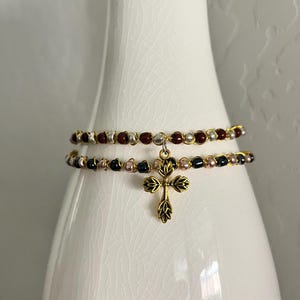 K&ouml;nnte beinhalten: Ein zweireihiges Perlenarmband mit einem goldfarbenen Kreuzanh&auml;nger in Blattform. Die obere Reihe besteht aus abwechselnd dunkelroten und silbernen Perlen, w&auml;hrend die untere Reihe abwechselnd schwarze und metallisch rosafarbene Perlen zeigt.