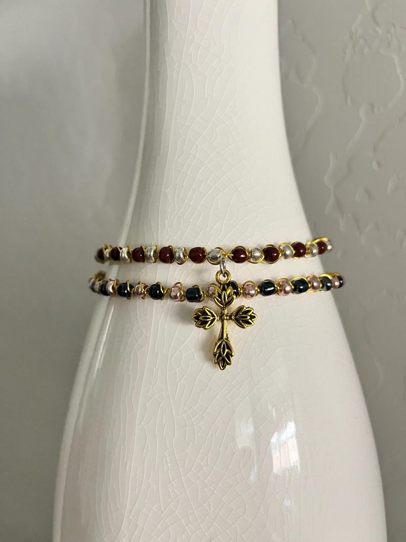 K&ouml;nnte beinhalten: Ein zweireihiges Perlenarmband mit einem goldfarbenen Kreuzanh&auml;nger in Blattform. Die obere Reihe besteht aus abwechselnd dunkelroten und silbernen Perlen, w&auml;hrend die untere Reihe abwechselnd schwarze und metallisch rosafarbene Perlen zeigt.