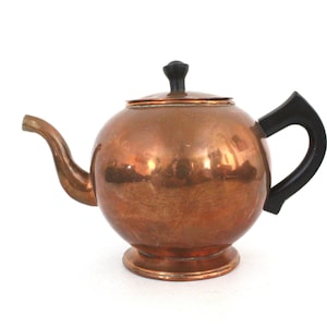 Puede incluir: Una tetera de cobre con asa y pomo de plástico negro. La tetera tiene un cuerpo redondo y una base acampanada.