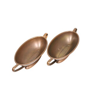 May include: Two oval-shaped metallic bowls with a warm bronze finish. Each bowl features a smooth, deep interior basin and two curved handles extending from opposite sides, providing a balanced and symmetrical appearance for these serving vessels.