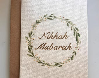 May include: A cream-colored card with the words "Nikkah Mubarak" in brown script, encircled by a green and gold floral wreath. The card is paired with a brown envelope, suitable for a wedding or celebratory occasion.