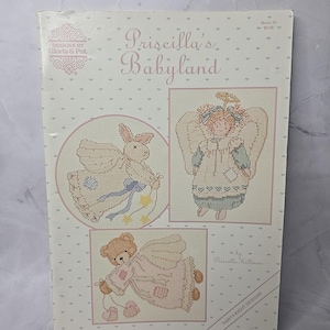 Peut inclure: Un livre de patrons de point de croix vintage intitulé "Priscilla's Babyland". La couverture présente trois motifs de point de croix : un ange, un lapin et un ours en peluche, tous avec des ailes. Le livre est conçu par Gloria & Pat et Priscilla Hillman.