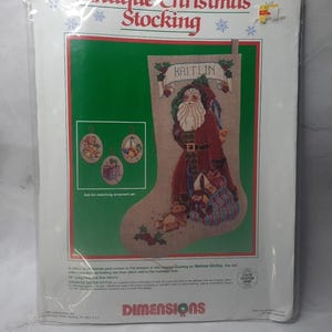May include: A cross stitch pattern for a Christmas stocking featuring a Santa Claus with a beard and a bag of gifts. The stocking is decorated with holly and the name "Kaitlin". The pattern includes three ornaments: a teddy bear, a sailboat, and a star. The pattern is from Dimensions and is called "Antique Christmas Stocking".