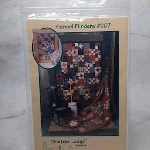 以下が含まれることがあります： パインツリーロッジデザインによる「フランネル・フリンダーズ＃207」というキルトのキルトパターンです。キルトは赤、白、青の生地でできており、星柄です。パターンは、ロッキングチェアと白いボールの入った瓶のあるテーブルの上に置かれています。
