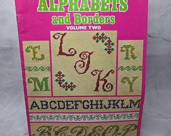 May include: A pink booklet titled "Charted Alphabets and Borders, Volume Two" with a leaflet number of 57 and a price of $2.00. The cover features a cross-stitch design of the letters L, J, K, M, and R.