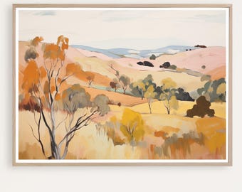 May include: A framed landscape art print featuring a warm color palette. The artwork depicts rolling hills and trees in shades of orange, yellow, and brown, with a light blue sky. The print is encased in a light-colored wooden frame.