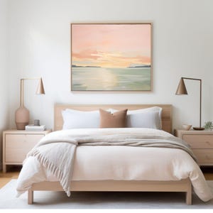 May include: A bedroom scene featuring a light wood bed frame with white bedding, a tan throw blanket, and a brown pillow. Two matching light wood nightstands flank the bed, each with a lamp. A large square painting of a sunset hangs above the bed.