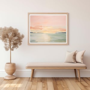 May include: A framed landscape painting with a serene sunset scene. The artwork features a soft color palette of pink, yellow, and blue, depicting a body of water and a distant shoreline. The frame is light-colored wood.