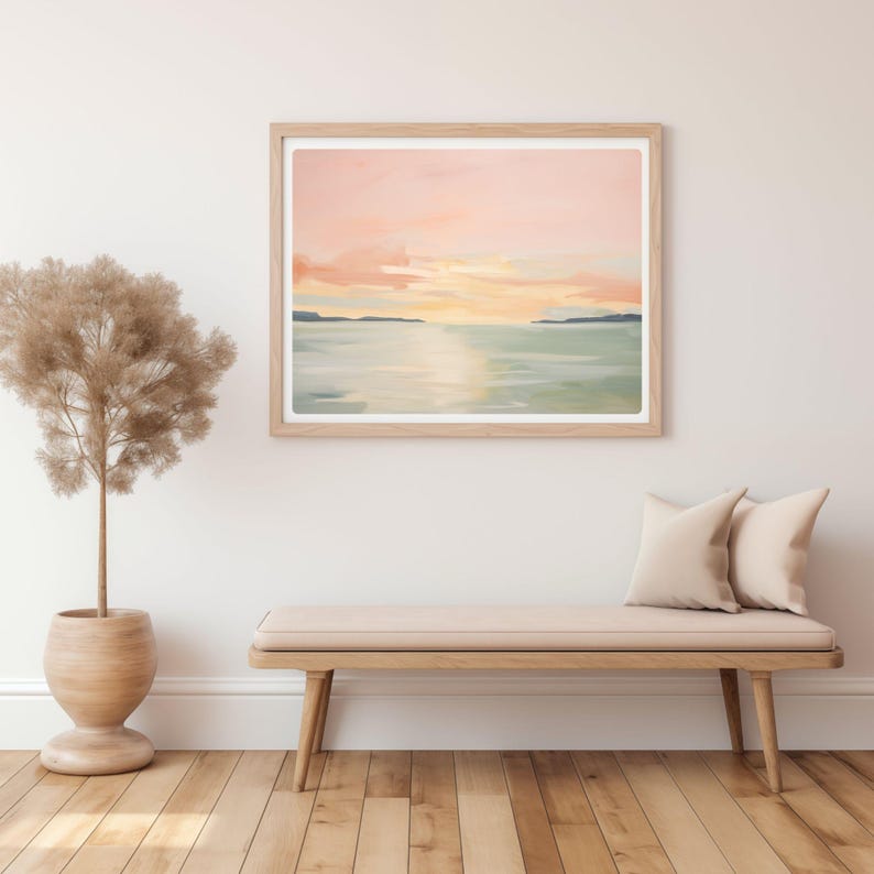 May include: A framed landscape painting with a serene sunset scene. The artwork features a soft color palette of pink, yellow, and blue, depicting a body of water and a distant shoreline. The frame is light-colored wood.