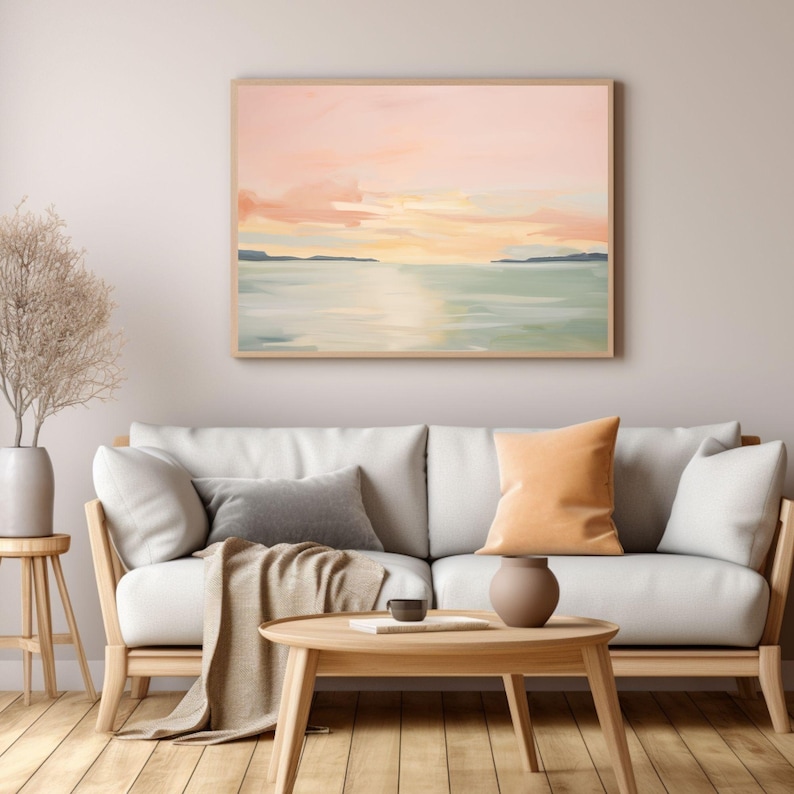 May include: Abstract landscape painting in a wooden frame. The artwork depicts a serene scene with a pastel sky, calm water, and distant land. The painting hangs above a light-colored sofa with pillows and a wooden coffee table.