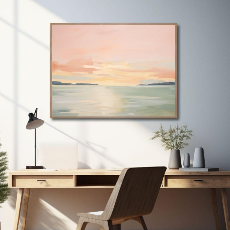 May include: A framed landscape painting depicts a serene sunset over water. The artwork features soft pastel colors of pink, orange, and green, with a wooden frame. The painting hangs above a wooden desk with a black lamp and a wooden chair.