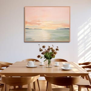 May include: An abstract landscape painting in a light wood frame hangs above a wooden dining table. The painting features a sunset with pink and orange hues over a body of water. A vase of dried flowers and a tea set are on the table.