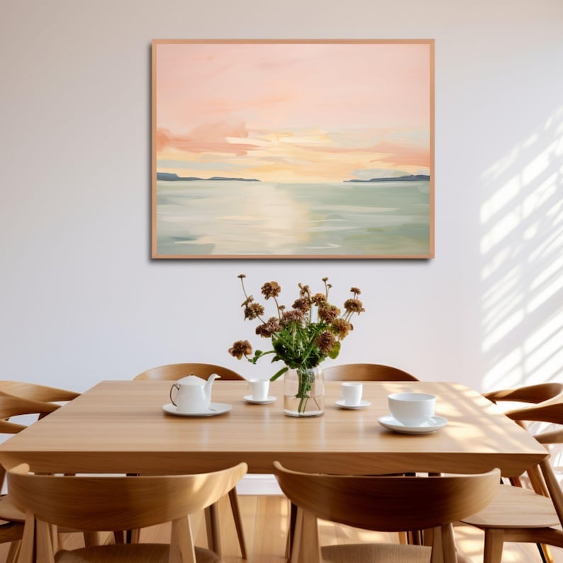 May include: An abstract landscape painting in a light wood frame hangs above a wooden dining table. The painting features a sunset with pink and orange hues over a body of water. A vase of dried flowers and a tea set are on the table.