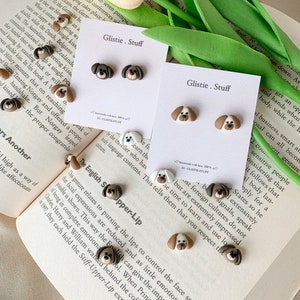 Może przedstawiać: Dwie karty z tekstem "Glistie. Stuff". Na każdej karcie znajdują się cztery małe kolczyki z polimerowej gliny w kształcie psiej głowy, w kolorze brązowym, białym i czarnym. Kolczyki są ułożone w siatkę na kartach.