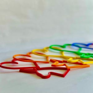 May include: A set of rainbow-colored plastic heart-shaped hoops. The hoops are arranged in a semi-circle, with the red hoop in the foreground and the blue hoop in the background.