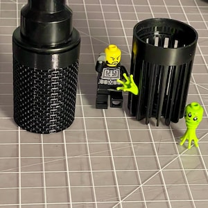 Può includere: Microfono in metallo nero con un design a rete, accanto a un oggetto cilindrico nero e due personaggi Lego. Un personaggio Lego è una minifigure in bianco e nero con testa gialla e mani verdi. L'altro è un alieno verde.