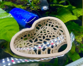 May include: A beige, heart-shaped aquarium decoration with a lattice design floats in the water. The decoration is surrounded by green aquatic plants and a blue cylindrical object. The aquarium is lit with white and red lights.