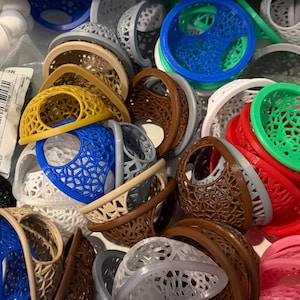 May include: A collection of 3D-printed, openwork baskets in various colors, including blue, yellow, brown, red, and green. The baskets have a geometric design and are stacked together. Some baskets are oval-shaped, while others are round.
