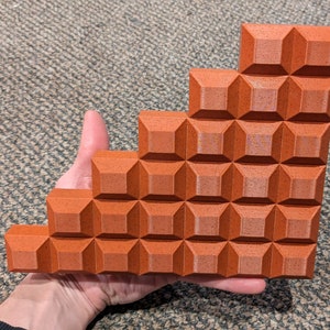 May include: A geometric, stepped brick-like object in a warm reddish-brown hue. The object is held in a hand and features a repeating pattern of square, raised blocks. The design resembles a miniature staircase or architectural model.