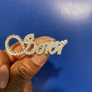 May include: Silver-toned "Sister" brooch with small, round, pearl-like embellishments. The brooch features a cursive font and is held against a blue background. The brooch is a jewelry item.