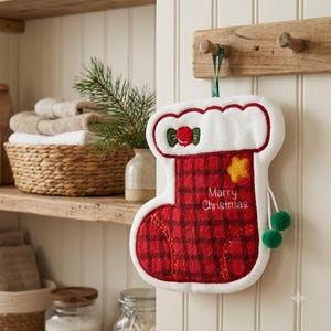 Könnte beinhalten: Ein rot-schwarz kariertes Handtuch in Form eines Weihnachtsstrumpfes mit weißem Rand. Das Handtuch zeigt einen gestickten gelben Stern, ein rotes Bonbon-Motiv und den Text "Marry Christmas" in Weiß. Es enthält eine grüne Aufhängeschlaufe und zwei grüne Bommeln.