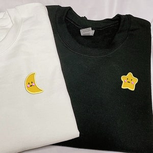 May include: Two white and black t-shirts with embroidered patches. The white t-shirt has a yellow crescent moon with a pink blush and a smiling face. The black t-shirt has a yellow star with a pink blush and a smiling face.