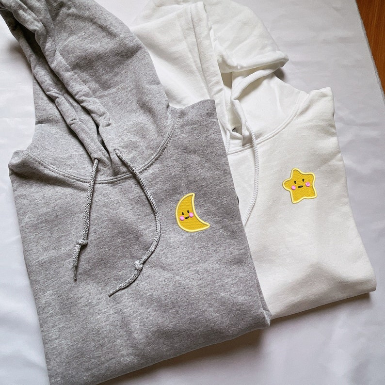 May include: Two hooded sweatshirts, one gray and one white, with embroidered cartoon faces. The gray sweatshirt has a yellow crescent moon with a smiling face, and the white sweatshirt has a yellow star with a smiling face.