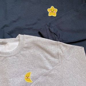 May include: Two sweatshirts with embroidered patches. The top sweatshirt is black with a yellow star patch with a pink face. The bottom sweatshirt is gray with a yellow crescent moon patch with a pink face.