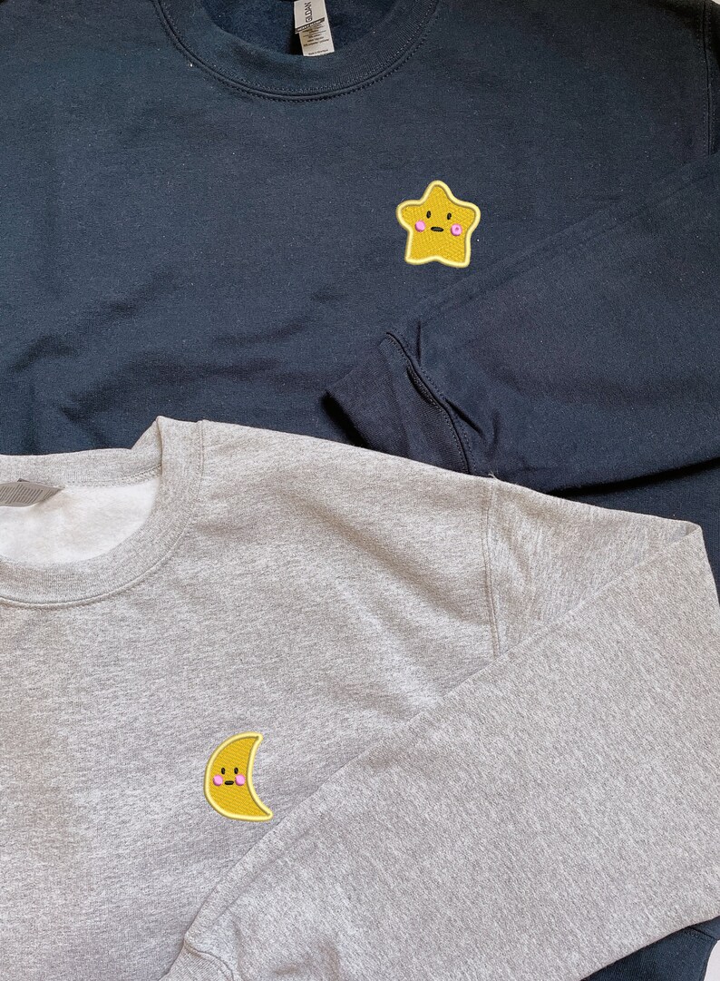 May include: Two sweatshirts with embroidered patches. The top sweatshirt is black with a yellow star patch with a pink face. The bottom sweatshirt is gray with a yellow crescent moon patch with a pink face.