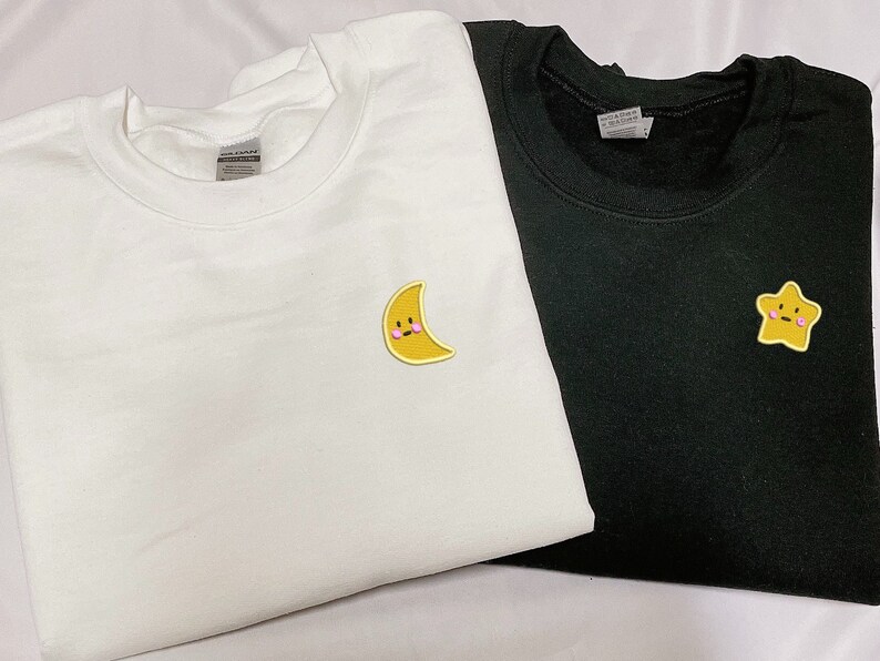 May include: Two white and black t-shirts with embroidered patches. The white t-shirt has a yellow crescent moon with a pink blush and a smiling face. The black t-shirt has a yellow star with a pink blush and a smiling face.