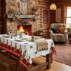 May include: A white rectangular tablecloth featuring a repeating pattern of nutcrackers and holly leaves, with a red and black plaid border. A tan burlap table runner with colorful tassels sits in the center of the table, which is set with plates.