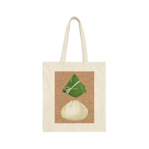 Puede incluir: Una bolsa de tela beige con un patrón marrón y blanco. La bolsa presenta una ilustración verde y blanca de una bola de masa y una ilustración verde y blanca de un pastel de arroz.