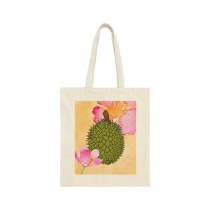 Puede incluir: Una bolsa de tela blanca con un diseño floral amarillo, rosa y naranja y un fruto de durian verde en el centro.
