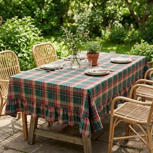 May include: A rectangular tablecloth featuring a green, red, and yellow plaid pattern with a ruffled edge. The tablecloth is draped over a wooden table set with white plates, clear glasses, a glass vase with flowers, and a potted plant.