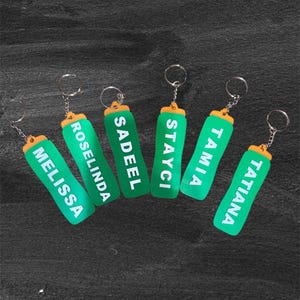 May include: Six green keychains with names in white lettering: MELISSA, ROSALINDA, SADEEL, STAYCI, TAMIA, and TATIANA. Each keychain has a silver ring and an orange top. The keychains are arranged on a dark gray surface.