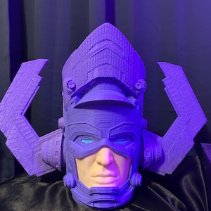 Puede incluir: Un casco morado con una cara, con un diseño detallado con elementos angulares y piezas laterales. La cara es de tono de piel claro con ojos azules. El casco es probablemente una pieza de disfraz o de colección.