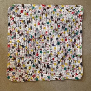May include: A square, handmade crochet blanket with a white base and colorful flecks of red, yellow, green, and brown. The blanket has a textured appearance and a white border.