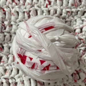 May include: A ball of white plastic shopping bags with red accents, tightly wound together. The ball is resting on a crocheted surface made from similar bags. The bags are knotted and woven, creating a textured, eco-friendly craft.