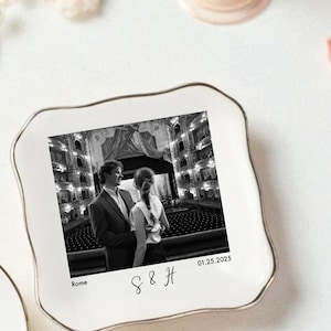 May include: Two white ceramic ring dishes with gold trim. One is heart-shaped, the other is square with scalloped edges. Both feature black and white photos of couples, with dates and initials. Decorative items.