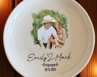 May include: A white ceramic plate with a painted image of a couple, with the text "Emily & Mark" and "Engaged 09.15.2025". The plate is surrounded by Christmas decorations.