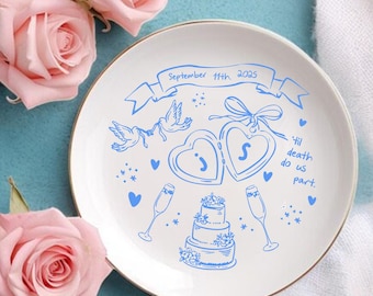 May include: A white ceramic plate with a gold rim, featuring blue illustrations of hearts, doves, a cake, and champagne flutes. The plate is decorated with the text "September 11th 2025" and "'til death do us part."