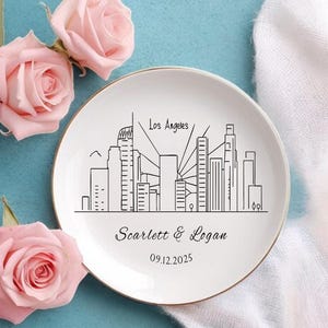 May include: A white ceramic plate with a gold rim features a black line drawing of the Los Angeles skyline. The plate is personalized with the names "Scarlett & Logan" and the date "09.12.2025". Pink roses are arranged around the plate.